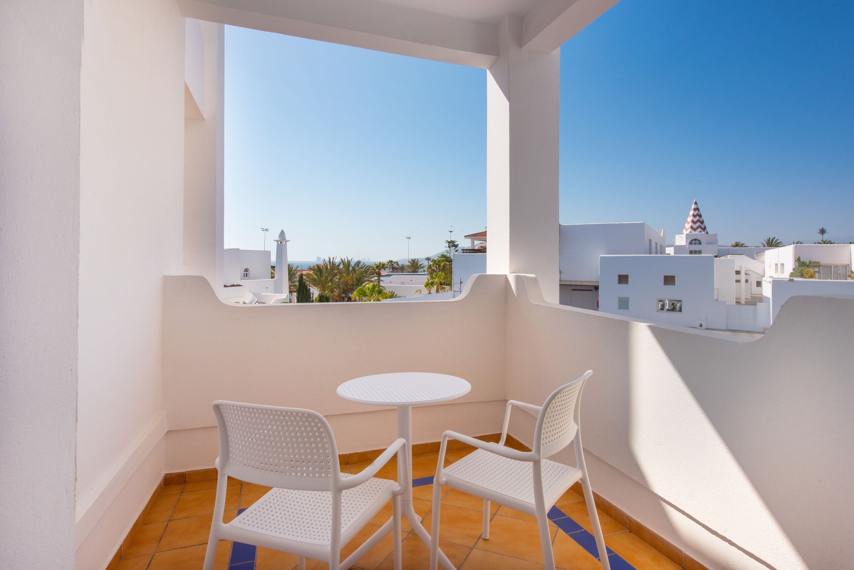 A balcony with white chairs and a small table, overlooking a cityscape with buildings and a clear blue sky.