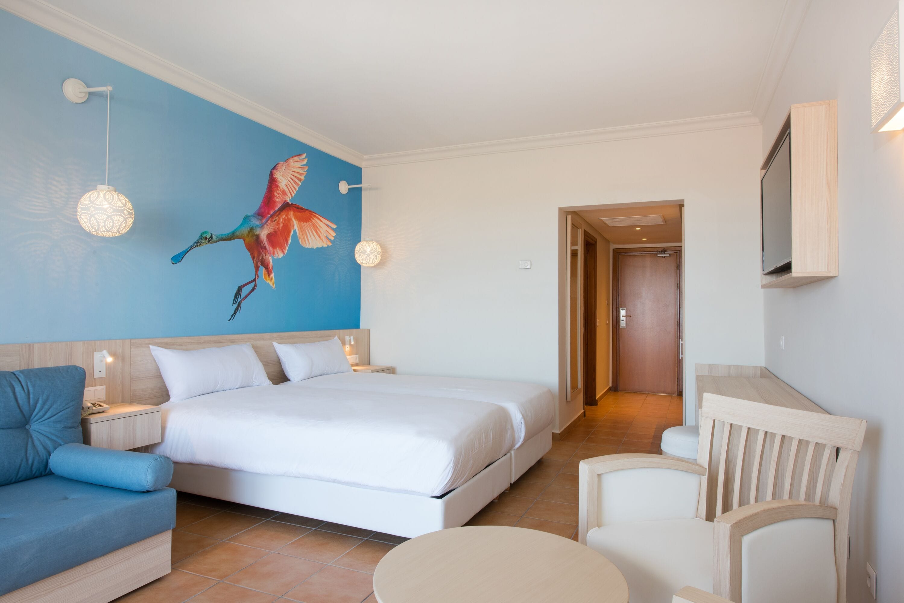 Ein Hotelzimmer mit einem großen Bett, einer blauen Wand mit Vogelgemälde, einem Fernseher und einem Holzstuhl.