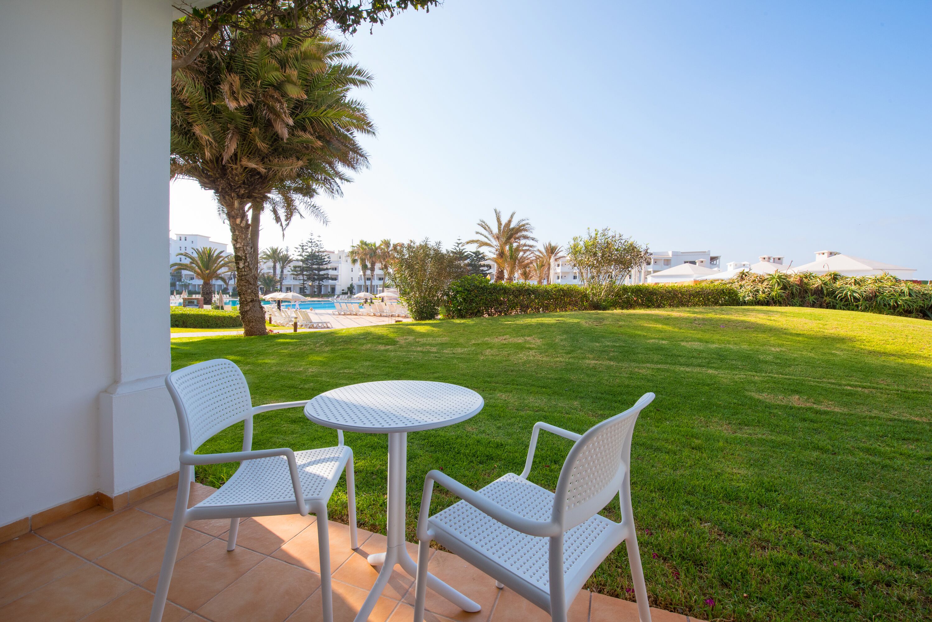 Une terrasse avec deux chaises blanches et une petite table ronde, donnant sur une pelouse bien entretenue et une zone piscine.