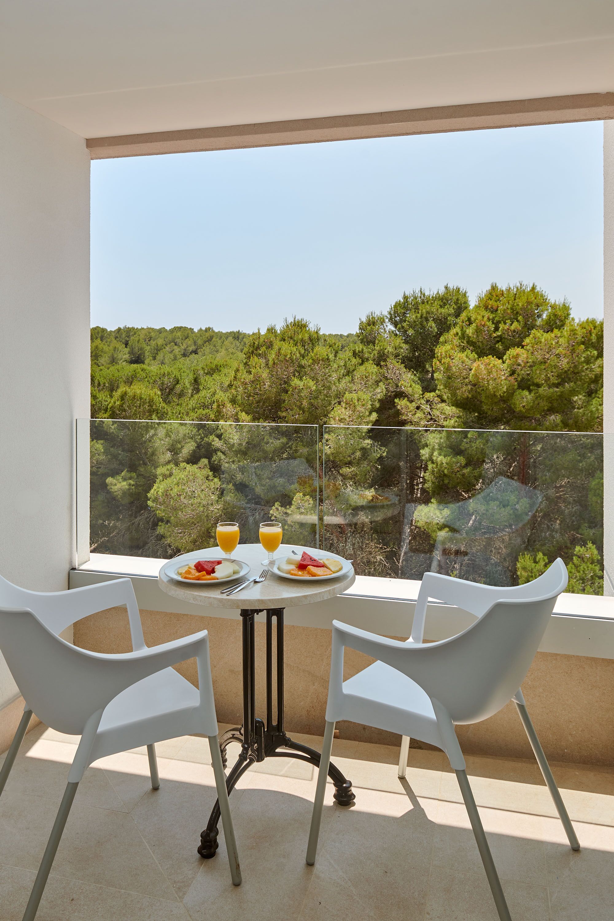 A balcony with two white chairs, a table with plates of food and drinks, and a view of a forested landscape.