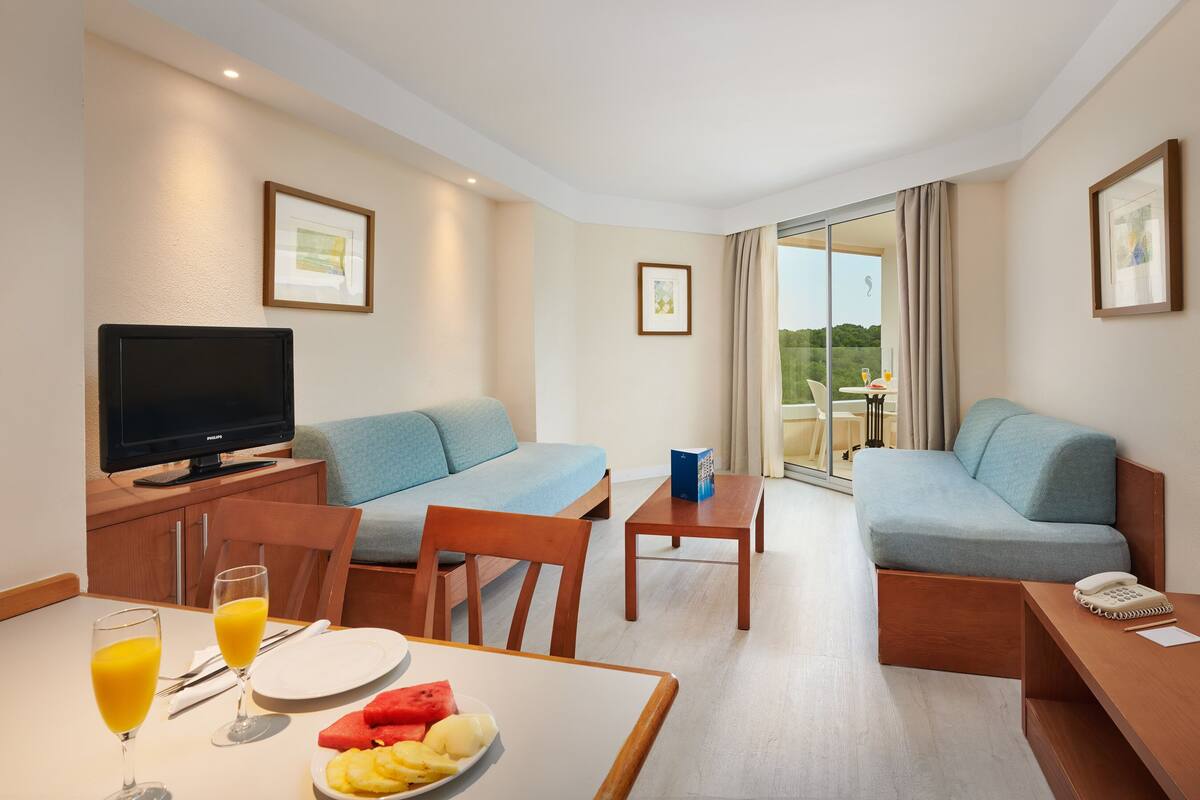 Ein Hotelzimmer mit Sofa, Fernseher, Esstisch mit Früchten und Saft sowie ein Balkon mit Tisch und Stühlen.