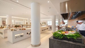 Un hall d’hôtel moderne avec un comptoir d’accueil central, de grandes colonnes blanches et un espace cuisine où un chef prépare les plats.