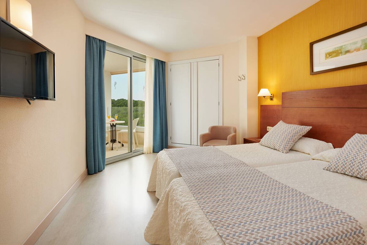Ein Hotelzimmer mit zwei Betten, einem Fernseher, einem Sessel und einem Balkon mit Tisch und Stühlen.