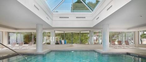 A modern indoor swimming pool with a skylight, clear glass walls, and lounge chairs.