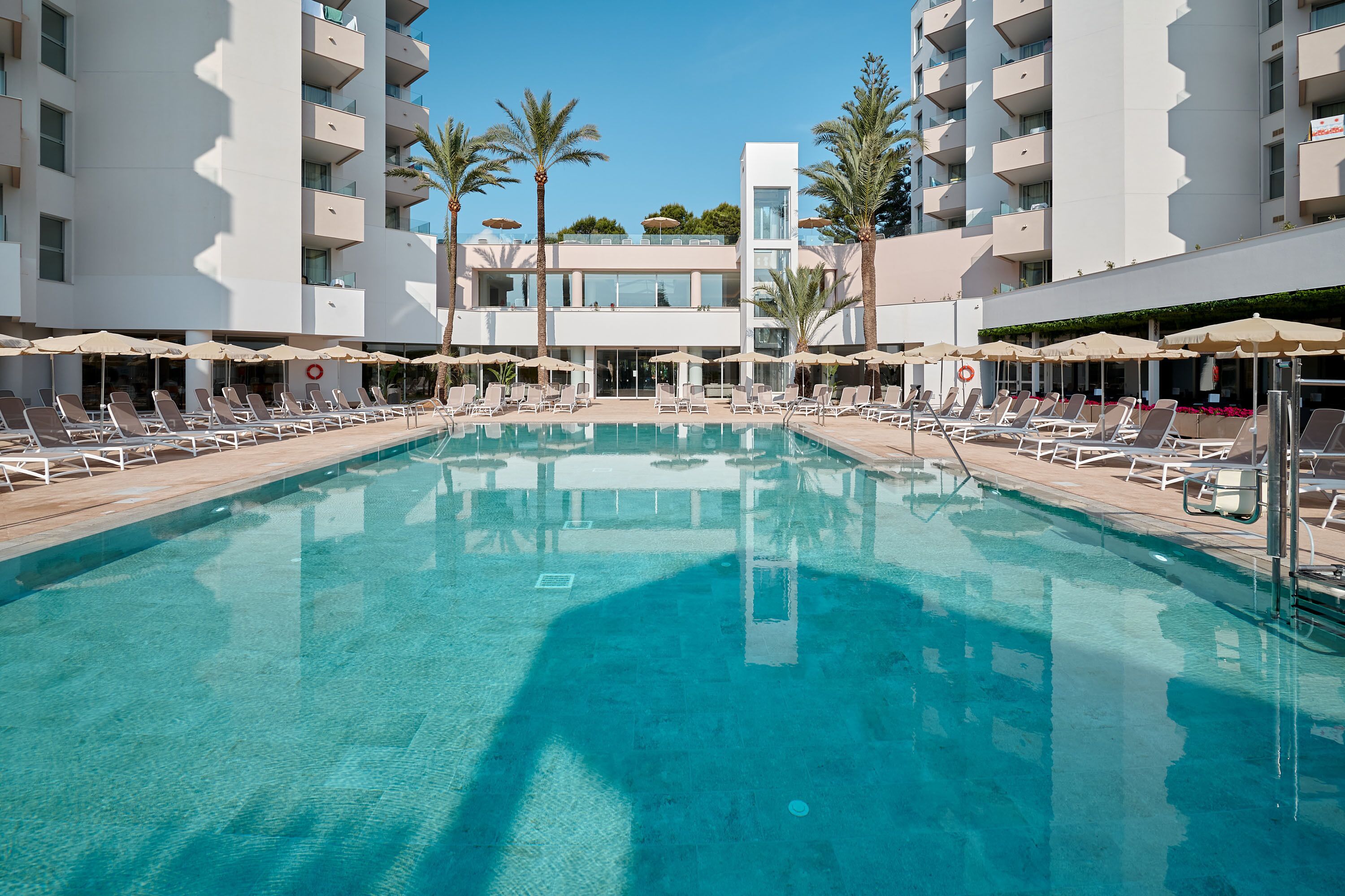 Ein Swimmingpool mit Liegestühlen und Sonnenschirmen, umgeben von einem modernen Gebäude mit Balkonen.