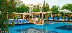 Una piscina con sillas de descanso y un área techada, rodeada de vegetación y edificios.
