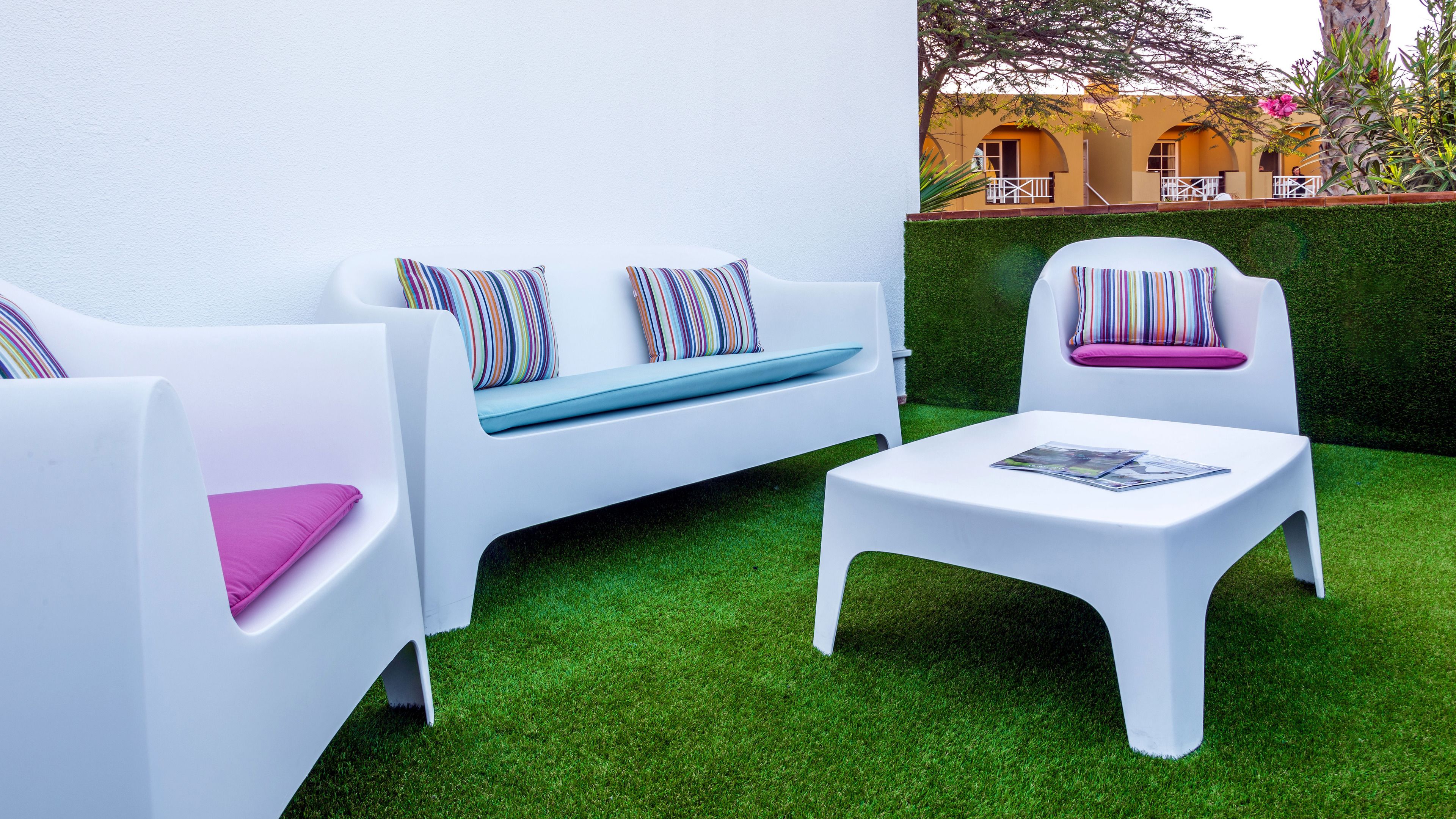 A modern outdoor seating area with white furniture and colorful cushions, surrounded by well-maintained green grass and a building with yellow walls in the background.