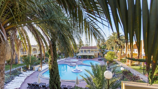 A pool area with lounge chairs and umbrellas, surrounded by palm trees and a building with a red roof. - Apartamentos Tisalaya Park (San Bartolome de Tirajana)