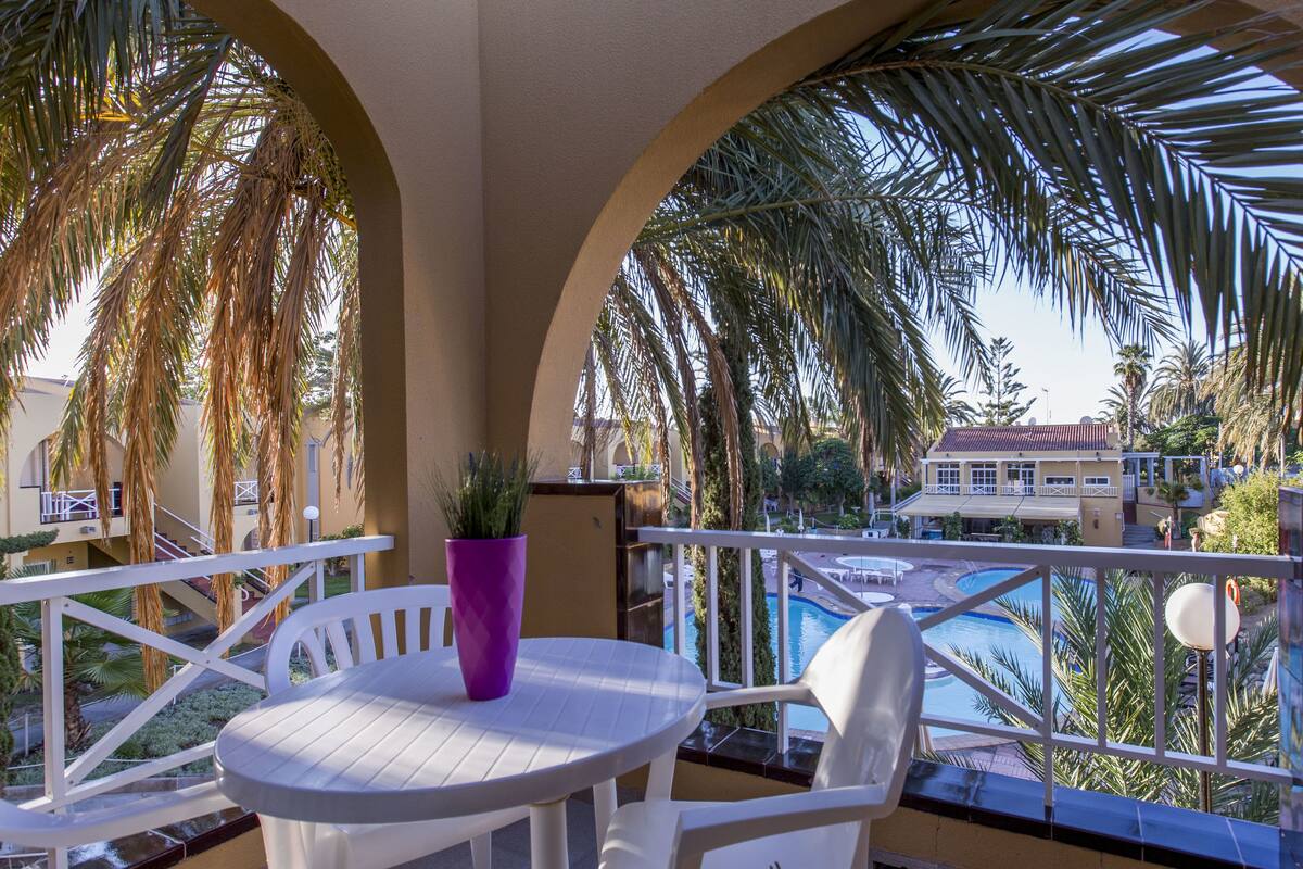 A balcony with a white table and chairs, a purple vase, and a view of a pool and buildings.