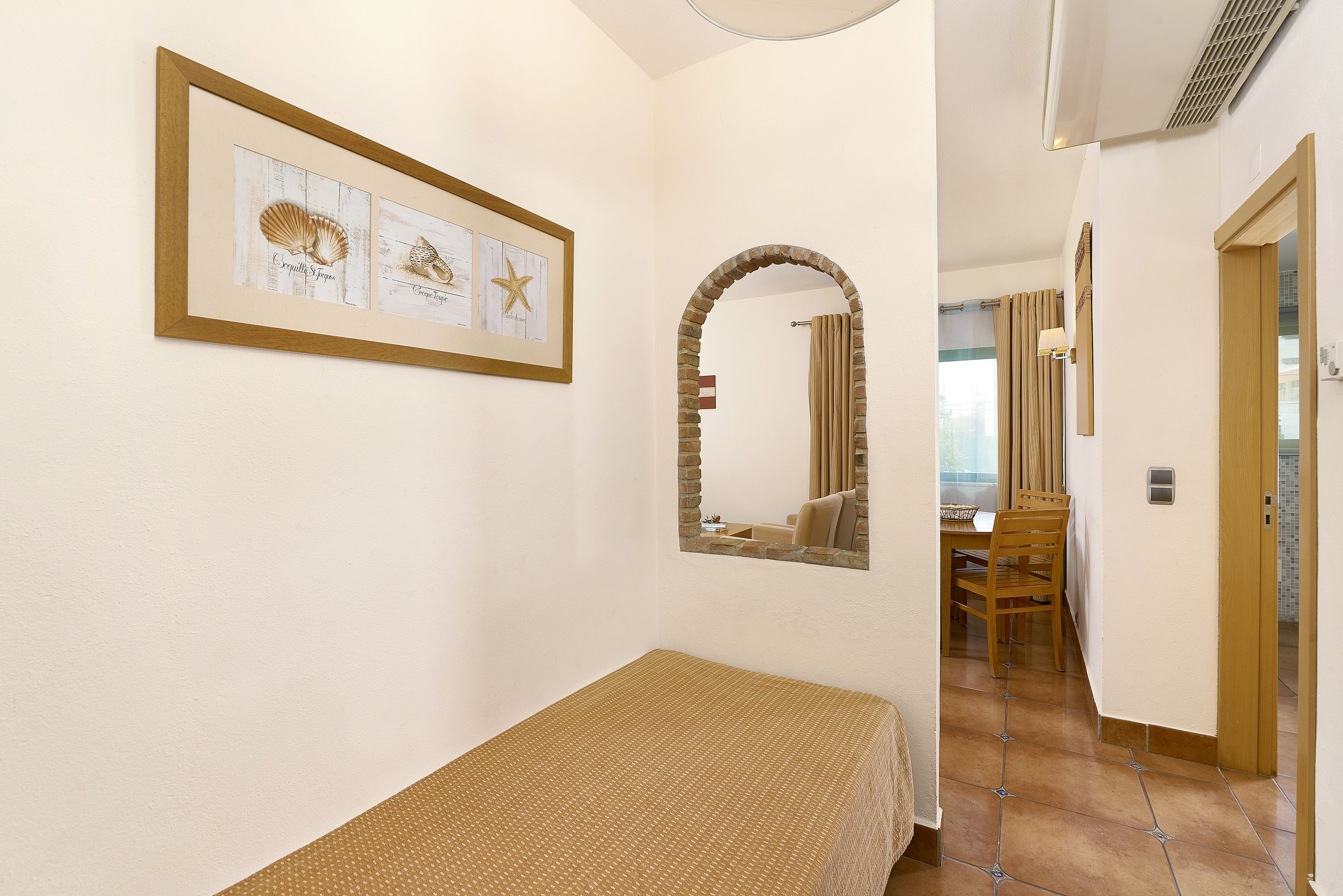 A bedroom with a bed, a framed picture of seashells, a mirror, and a view into the dining area.