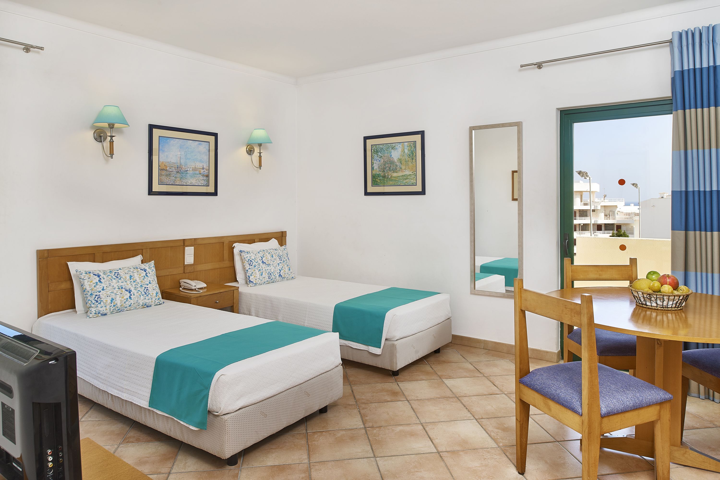 A hotel room with two beds, a desk with a chair, a TV, and a dining table with a fruit basket.