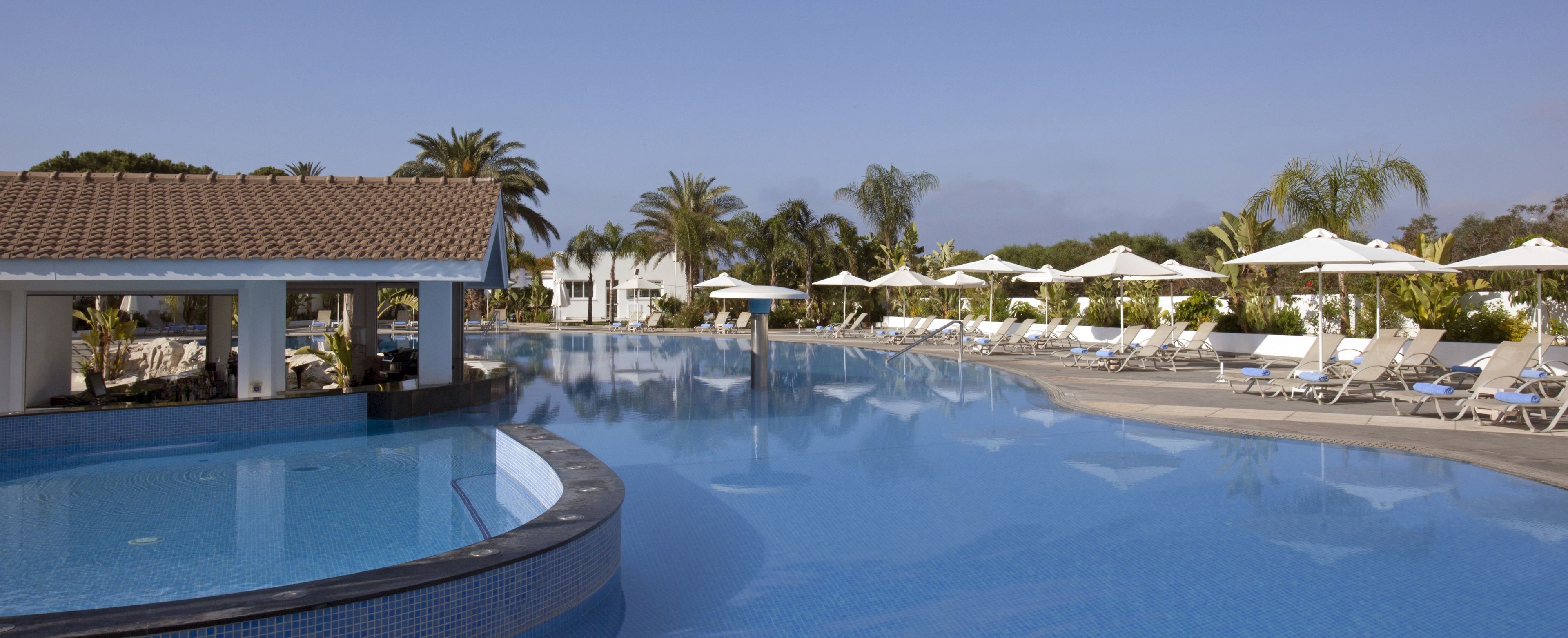 A swimming pool with lounge chairs and umbrellas, surrounded by palm trees and a building with a tiled roof.