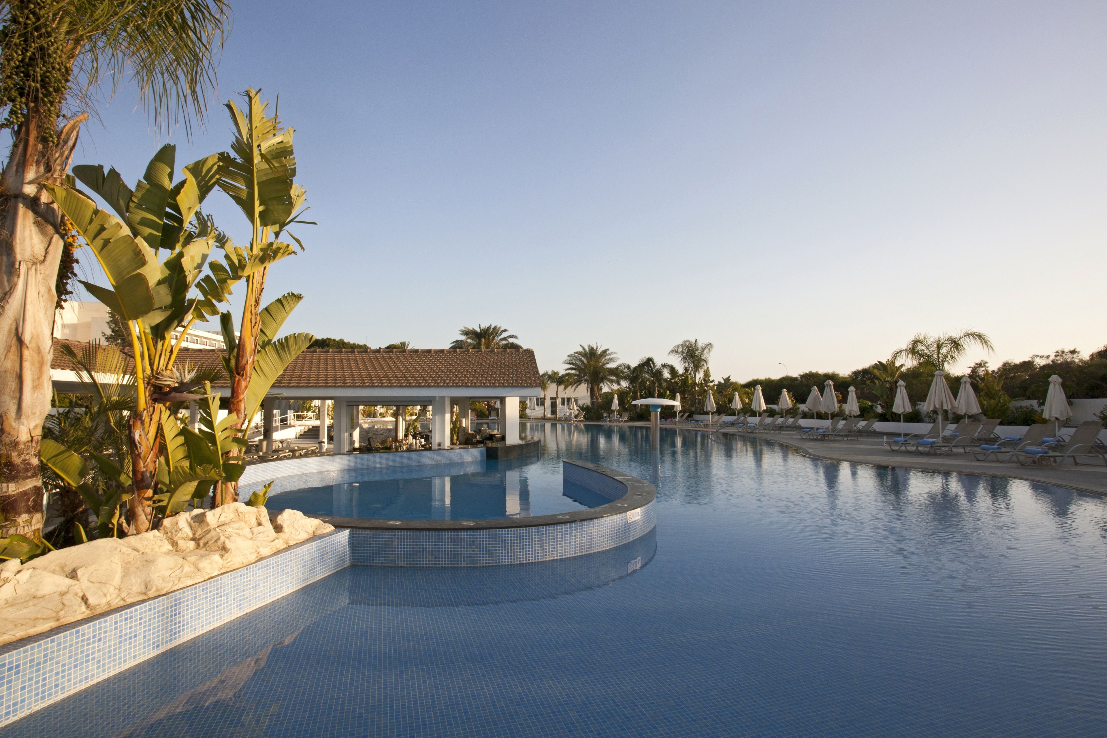 A swimming pool with lounge chairs and umbrellas, surrounded by palm trees and a building with a tiled roof.