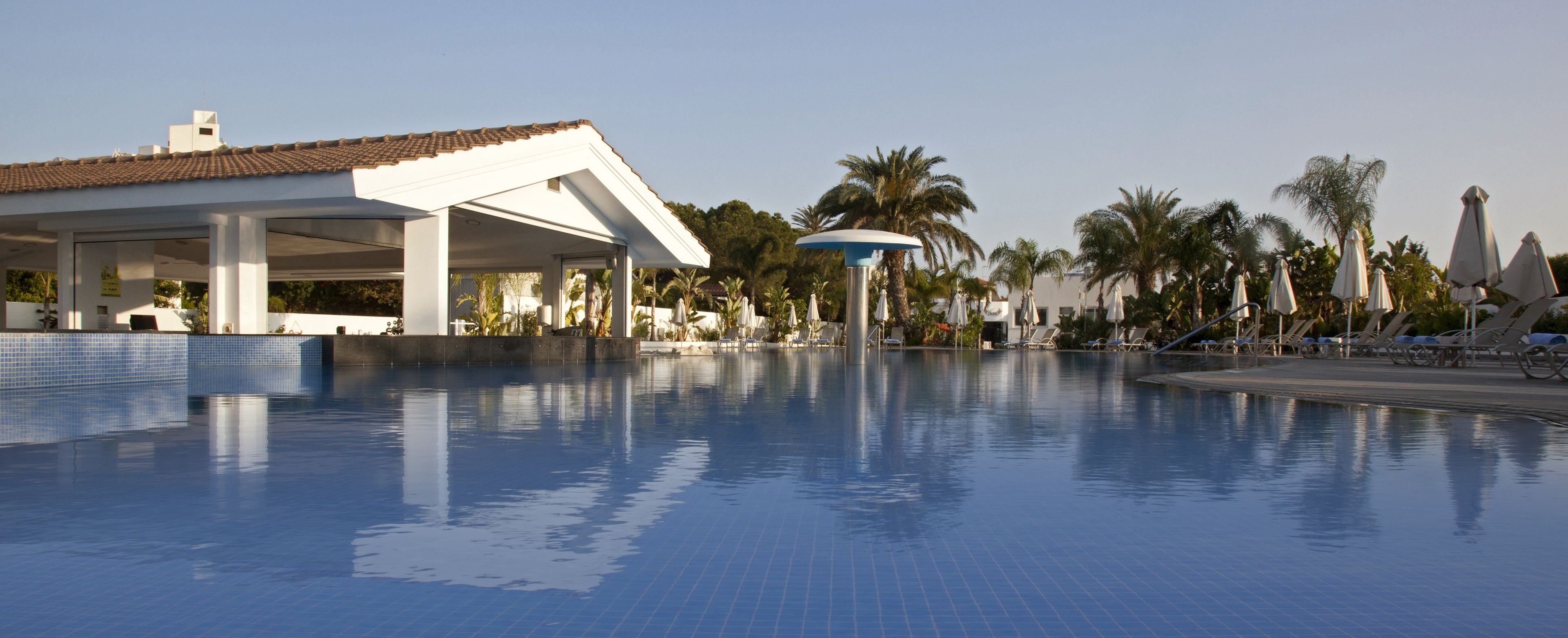 A swimming pool with lounge chairs and umbrellas, surrounded by palm trees and a building with a tiled roof.