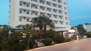 A multi-story hotel with balconies, a covered entrance, and a palm tree in the foreground. - Minay (Kusadasi)