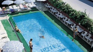A rectangular swimming pool with a green deck, surrounded by white lounge chairs and umbrellas. - Minay (Kusadasi)