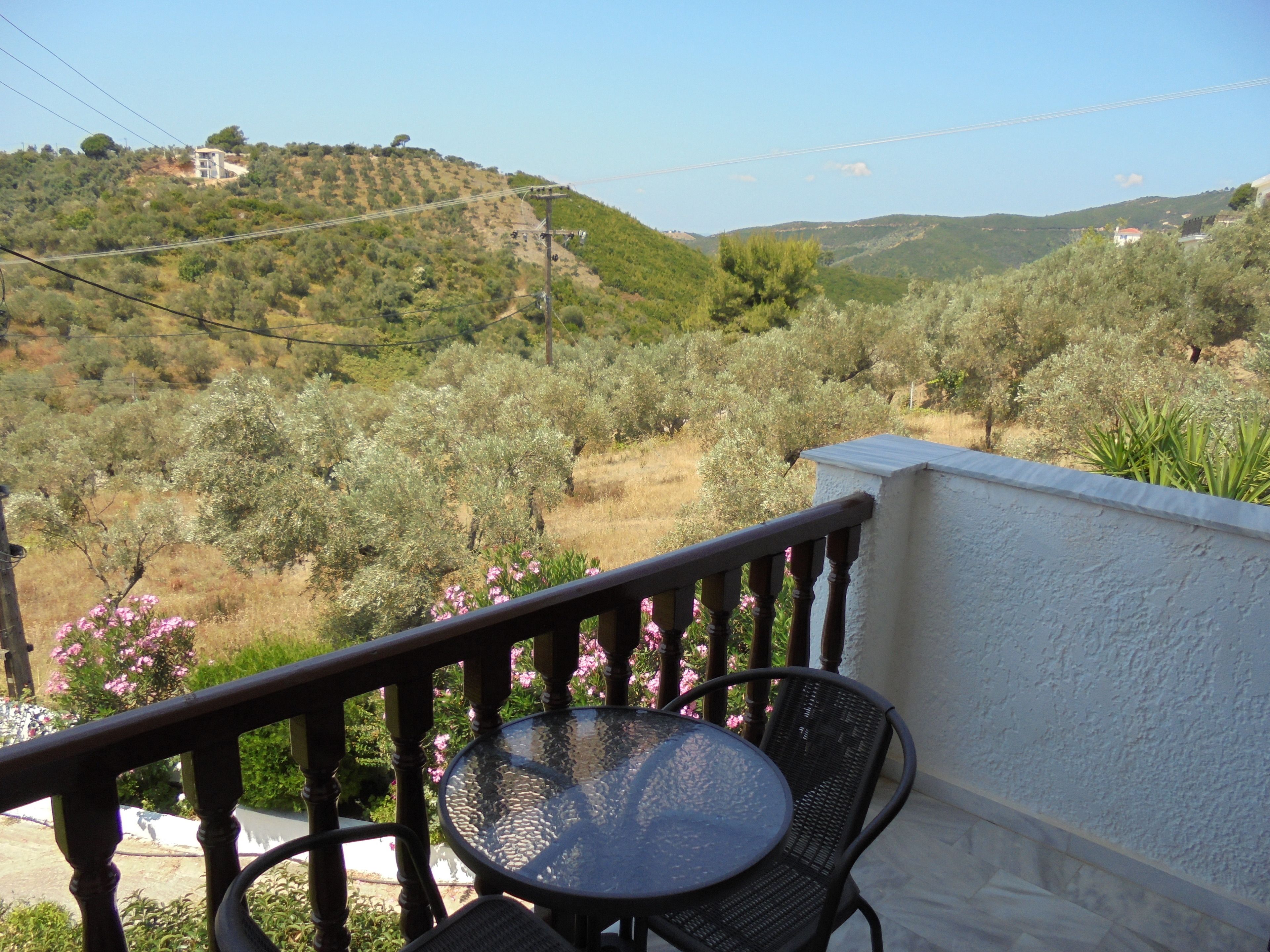 A balcony with a round table and two chairs, overlooking a hilly landscape with olive trees and a distant building.
