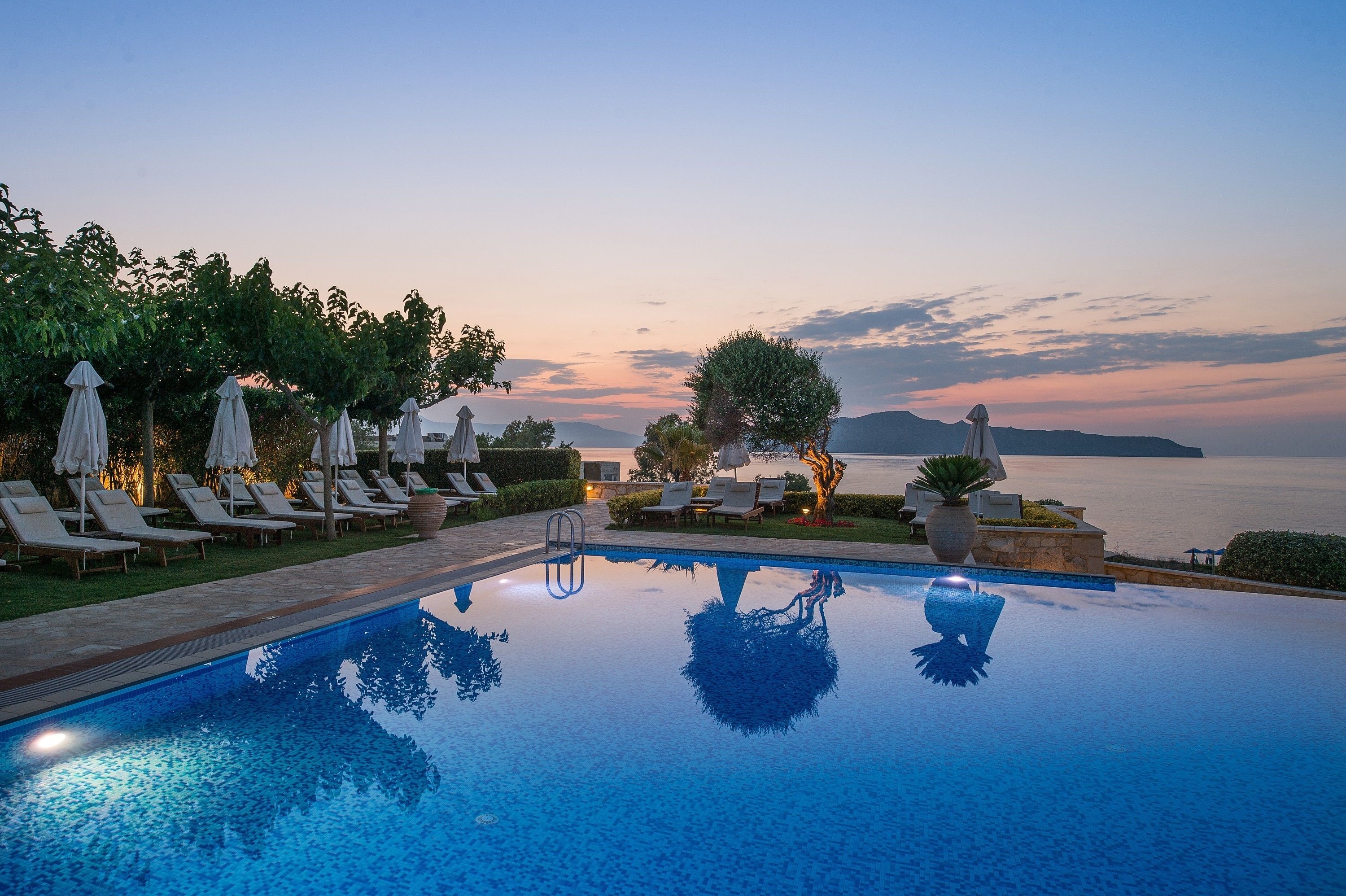 A serene pool area with lounge chairs and umbrellas, overlooking a calm body of water at dusk.