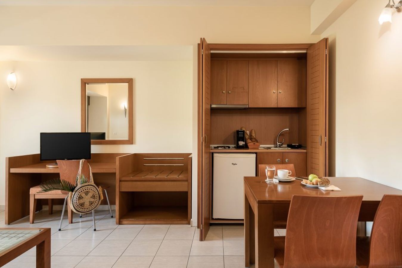 A compact hotel room with a kitchenette, a desk with a computer, a dining table, and a chair.