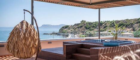 A sunlit outdoor seating area with a wicker chair, a small pool, and a view of the sea and hills.