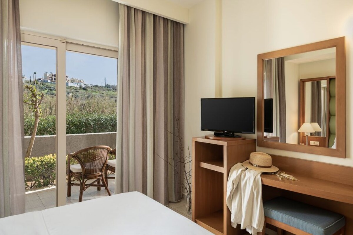 Habitación de hotel con una cama grande, un televisor sobre un soporte de madera, una silla, un espejo y un balcón con cortinas.