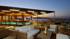 Un área de estar al aire libre con mobiliario de mimbre, una barra con mostrador y vistas al mar al atardecer.