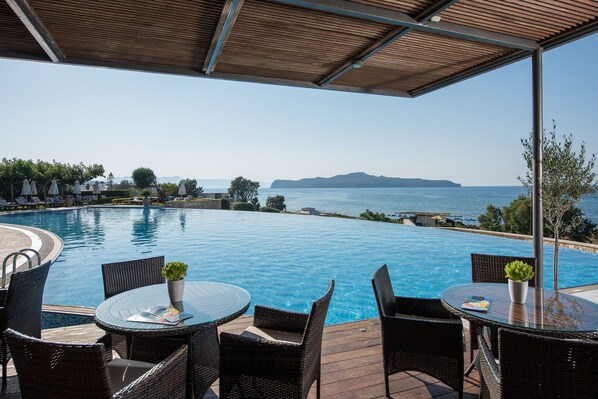 Zona junto a la piscina con mobiliario de mimbre y una mesa de vidrio, con vistas a una piscina azul cristalina y una lejana panorámica del mar y las montañas.