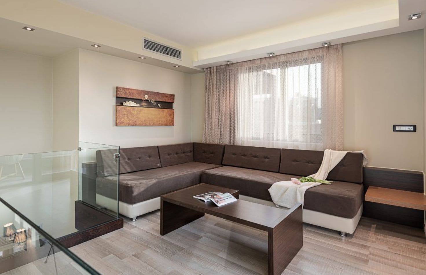 Een moderne woonkamer met een modulaire bank, een glazen salontafel en een groot raam met gordijnen.
