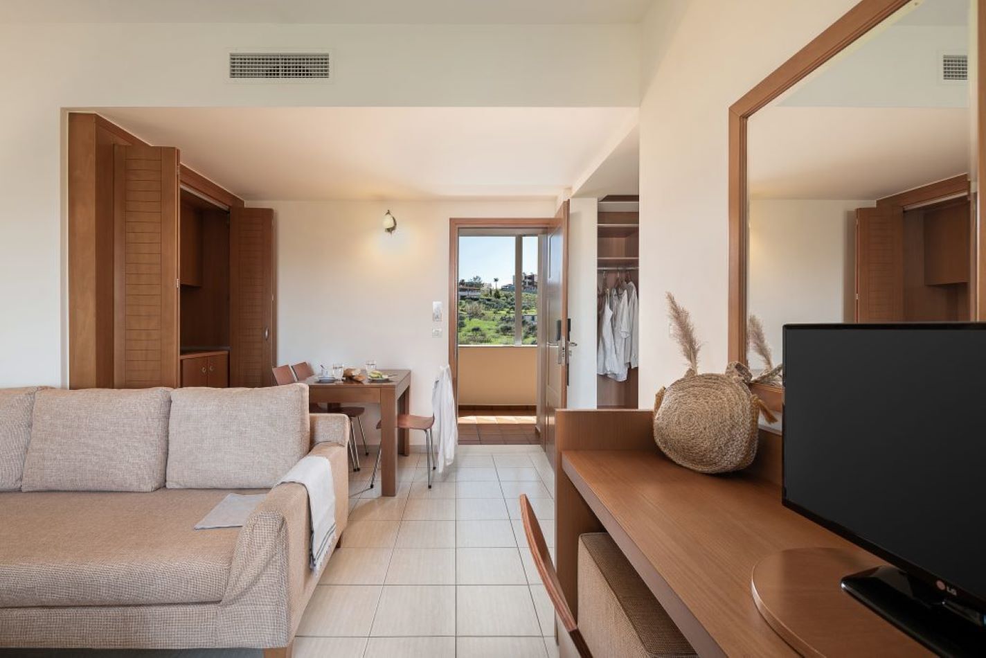 A modern living room with a sofa, a dining table, and a TV. There is a balcony with a view of the hills.