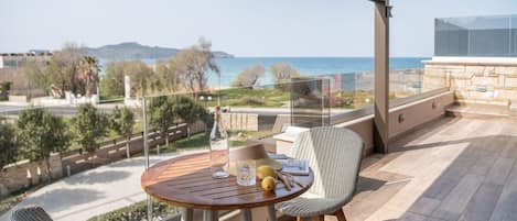 A balcony with a round wooden table, wicker chairs, and a clear view of the sea and distant hills.