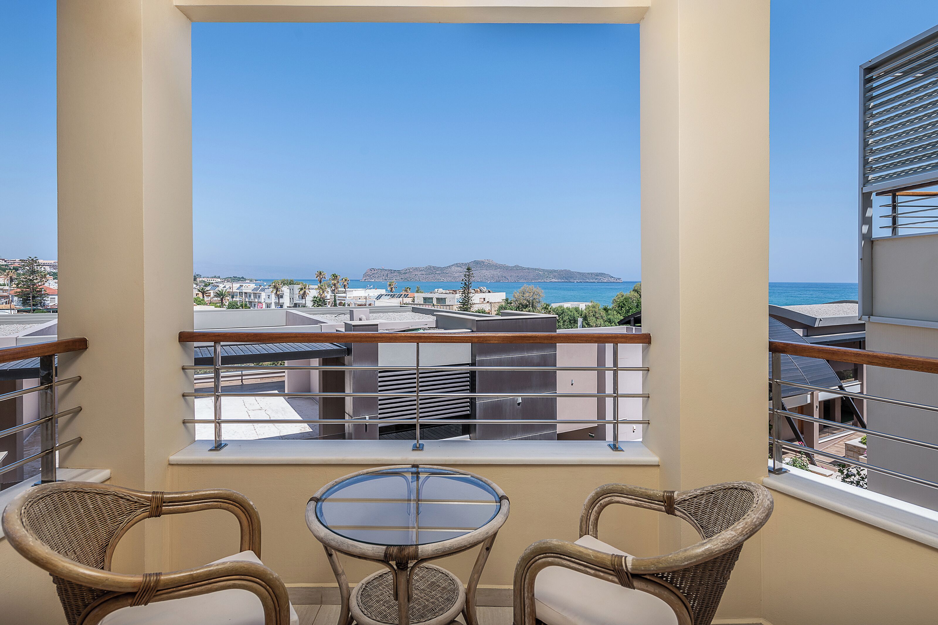 Un balcon agrémenté de fauteuils en osier et d’une table en verre, offrant une vue sur un paysage urbain côtier avec ses bâtiments et une île lointaine.