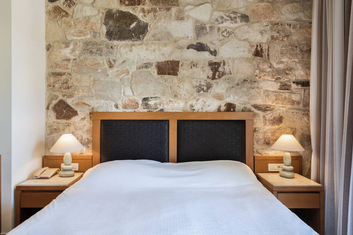 Hotellihuone, jossa on kiviseinä, valkoisilla lakanoiden peitetty sänky, kaksi yöpöytää, joissa on lamput, ja puhelin toisella yöpöydällä.