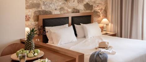 A hotel room with a stone wall, a bed with white linens, a wooden nightstand, a table with fruit and a glass, and a hat on the bed.