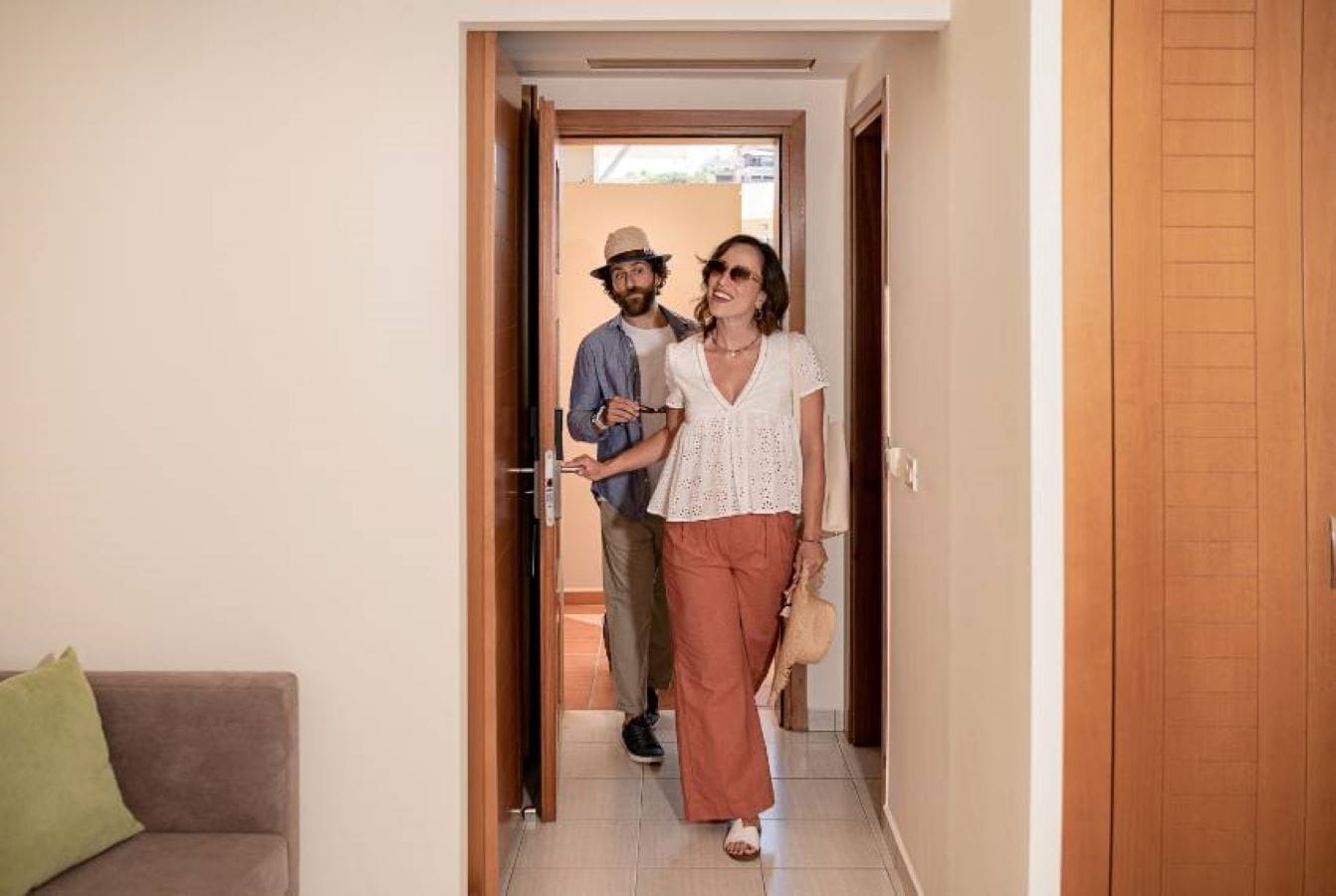 A hallway with a wooden door, a brown sofa with a green pillow, and a person holding a handbag.