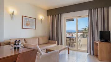 A modern living room with a sofa, coffee table, and dining area. There is a TV on a wooden stand and a balcony with a view.