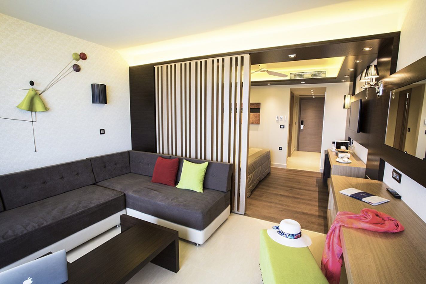 A modern hotel room with a sectional sofa, a desk with a laptop, a dining area, and a separate sleeping area.