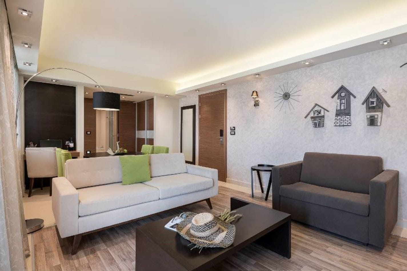 Een moderne woonkamer met een witte bank, een donkerne fauteuil en een salontafel. Er is een bureau met een computer en een lamp. De kamer heeft een houten vloer en een wand met decoratieve details.