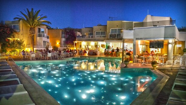A pool area with lounge chairs and tables, surrounded by buildings with outdoor seating and a palm tree. - Nefeli Hotel (Rethymno)