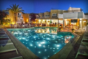 A pool area with lounge chairs and tables, surrounded by buildings with outdoor seating and a palm tree. - Nefeli Hotel (Rethymno)
