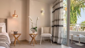 A modern hotel room with a bed, two small wooden tables, a white chair, and a balcony with a view of palm trees. - Nefeli Hotel (Rethymno)