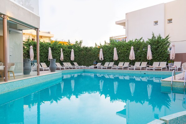 A swimming pool with lounge chairs and umbrellas, surrounded by greenery and modern buildings. - Nefeli Hotel (Rethymno)