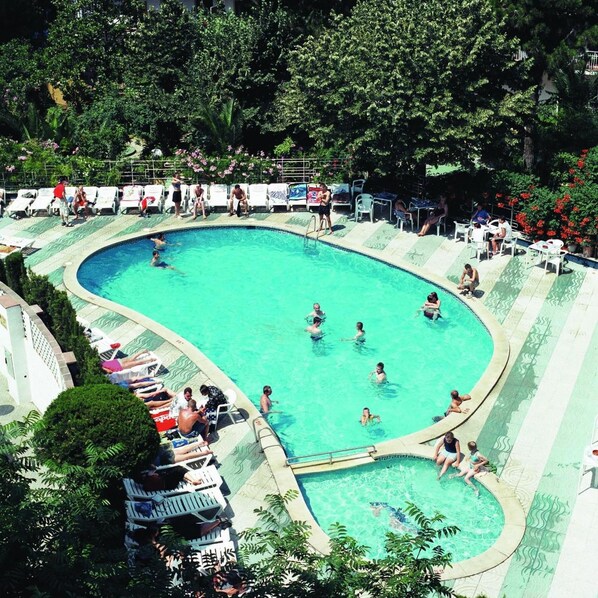 A swimming pool with multiple lanes, surrounded by lounge chairs and tables, set in a lush green environment. - Hotel Clipper (Lloret de Mar)