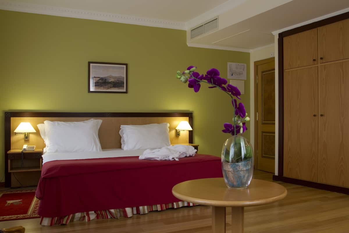 Una habitación de hotel con una cama, dos lámparas de noche, un florero con flores y un armario de madera.