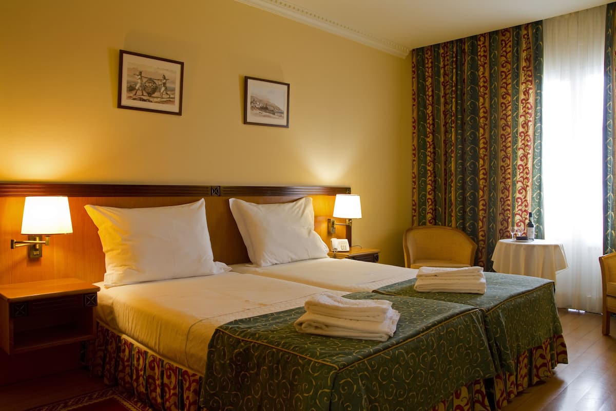 Habitación de hotel con dos camas, una mesita de noche con una lámpara, una silla, una pequeña mesa con una botella y dos cuadros en la pared.