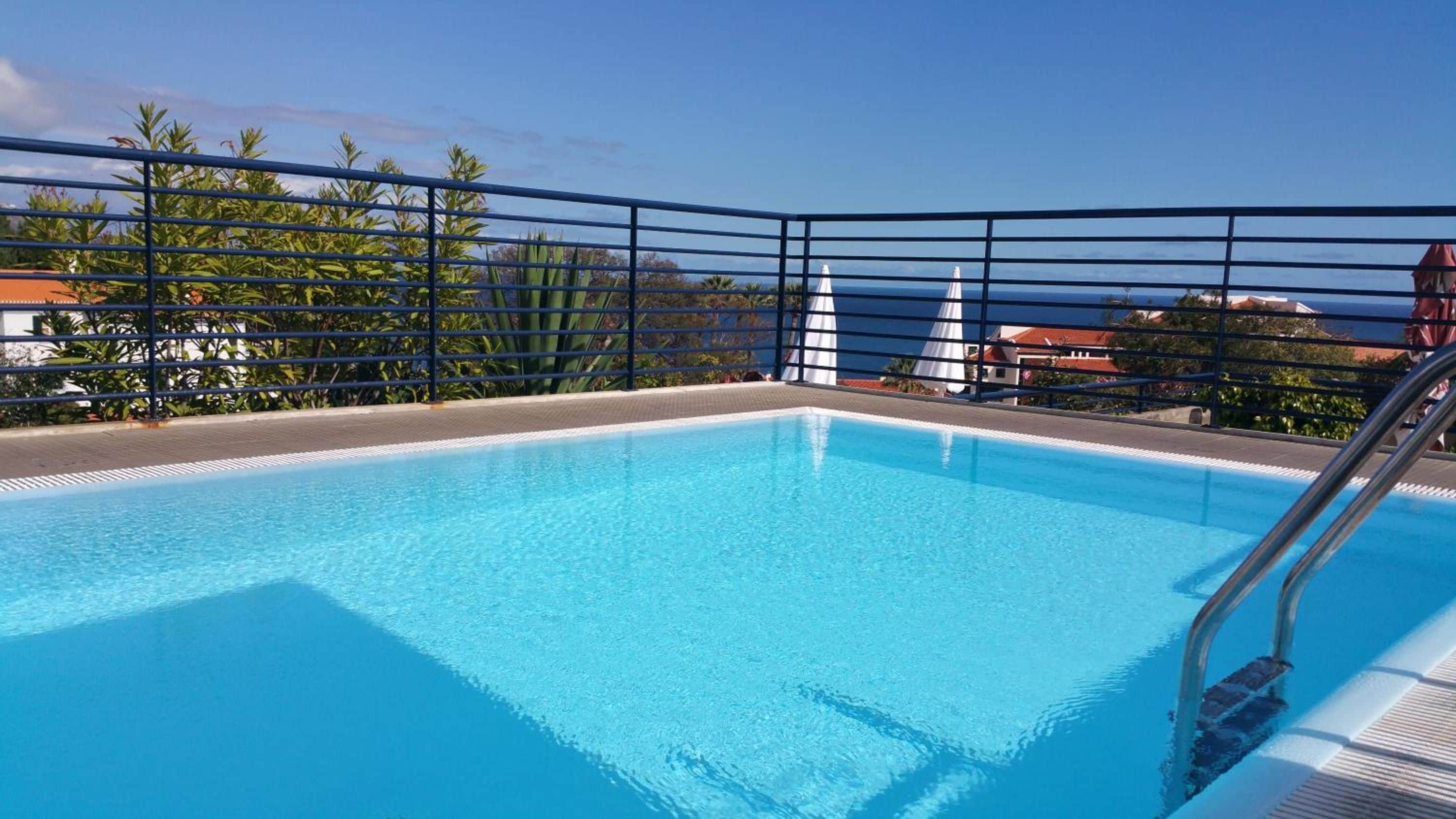 Piscina azul cristalina con barandilla metálica, rodeada de vegetación y vistas al mar.