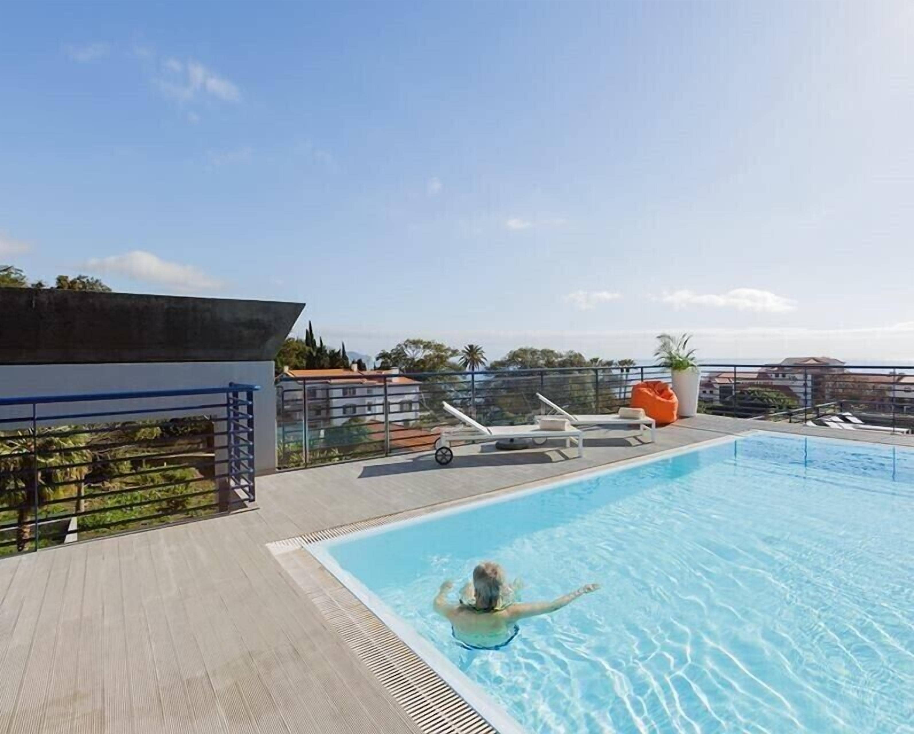 A rooftop swimming pool with a person swimming, surrounded by lounge chairs and a view of buildings and greenery.