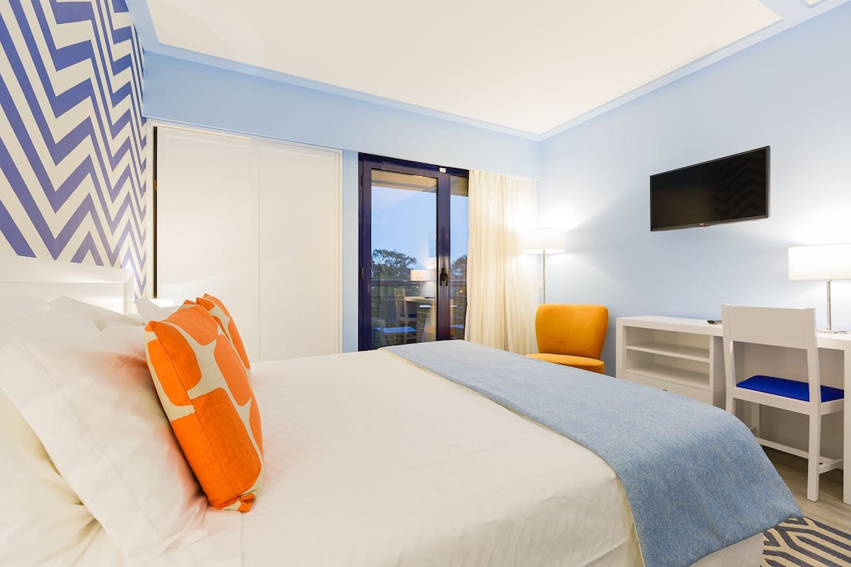 A hotel room with a bed, a desk, a chair, a TV, and a view through a glass door.