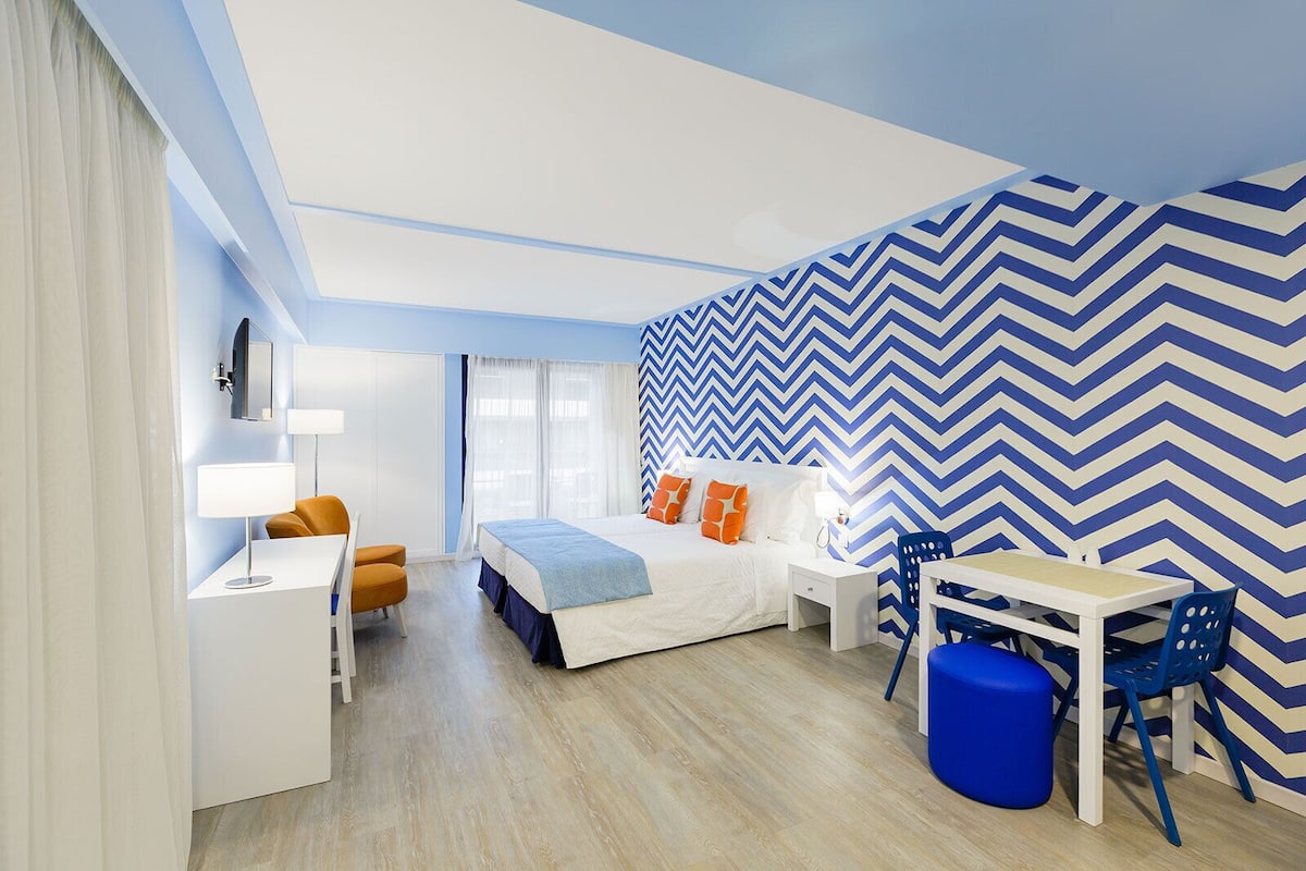 A modern hotel room with a bed, desk, chair, and a large window.