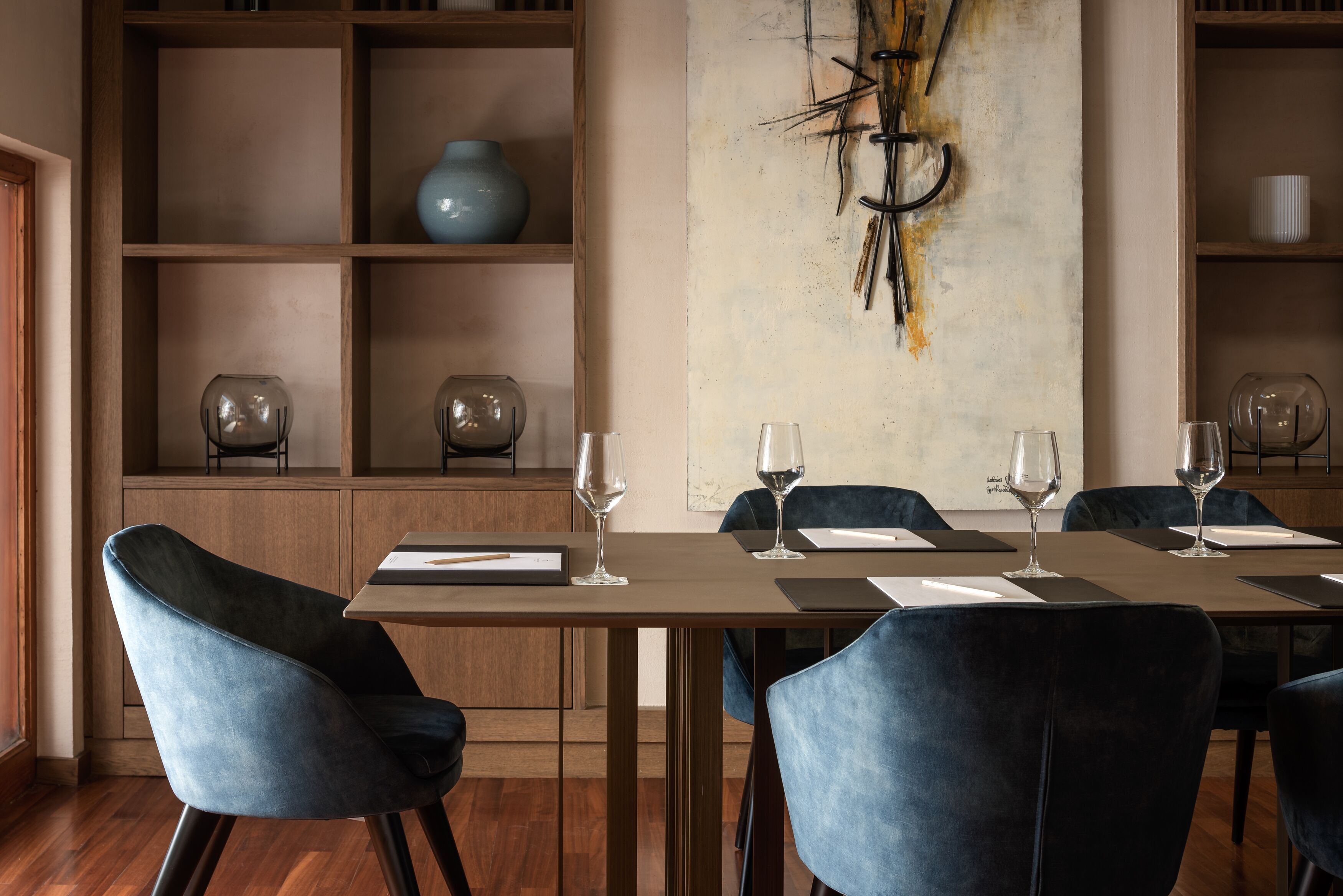 Un comedor con una mesa larga, sillas azules y copas de vino. Se observan estantes con objetos decorativos y un cuadro abstracto de gran tamaño en la pared.