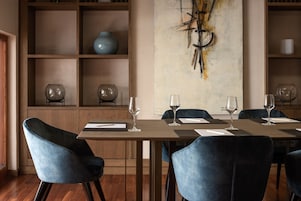 Un comedor con una mesa larga, sillas azules y copas de vino. Se observan estantes con objetos decorativos y un cuadro abstracto de gran tamaño en la pared.